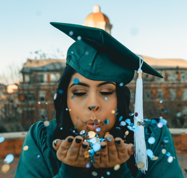 Graduating student in cap and gown blowing confetti